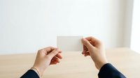 Young adult with medium skin tone holding blank white business card over wooden table, wearing dark sleeve, suitable for corporate branding, business identity, and professional marketing materials.