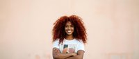 Happy young African American woman with curly hair smiling confidently with arms crossed against neutral beige wall background for lifestyle content.