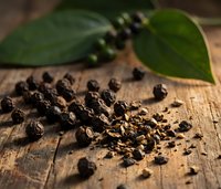 Black peppercorns scattered on rustic wooden surface with fresh green pepper leaves for culinary seasoning and spice cooking applications.