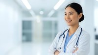 Confident Asian female doctor smiling with crossed arms in modern hospital corridor. Professional healthcare worker in white coat with stethoscope.