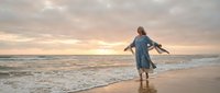 Young woman in blue dress walking barefoot on ocean beach at golden sunset with arms outstretched feeling freedom and joy during peaceful evening moment.