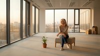 Young woman sitting thoughtfully in modern empty office space with large windows overlooking city skyline during golden hour sunset light.