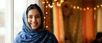 Young Middle Eastern woman wearing traditional blue hijab headscarf smiling warmly indoors with festive bokeh lights background for cultural diversity content.