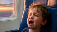 Toddler boy with curly brown hair crying and upset in airplane seat during flight with sunset visible through window.