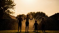 Children playing in sprinkler water during golden hour sunset creating magical silhouettes and refreshing summer fun in backyard.