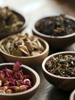 Assorted dried herbs and flowers in wooden bowls, including rose buds and loose tea leaves, arranged on dark surface with soft natural lighting.