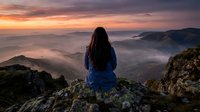 Woman with long dark hair wearing blue jacket sitting on rocky mountain peak overlooking misty valley at sunrise, contemplative travel and nature scene.
