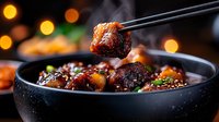 Asian style glazed meatballs with sesame seeds and green onions in black ceramic bowl. Chopsticks holding meatball against blurred bokeh lights background.