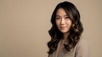 Professional Asian businesswoman with wavy hair in beige blazer smiling confidently against neutral background for corporate marketing materials.