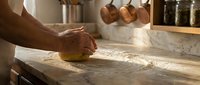 Professional baker kneading fresh dough on floured marble counter in rustic kitchen with copper pots and spice jars for artisan bread making.