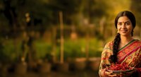Young South Asian woman wearing traditional red and gold patterned saree in lush garden setting with soft natural lighting and blurred foliage background.