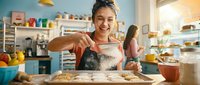 Happy young Asian woman baker decorating cookies with icing in bright modern kitchen, smiling while working on homemade treats for bakery business.