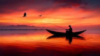 Silhouette of fisherman in traditional boat against dramatic orange sunset sky with flying birds and mountain backdrop reflecting on calm water surface.