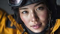 Close-up portrait of Asian female skier wearing goggles and yellow jacket, with snow flakes on face against dark background.