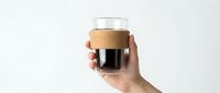 Hand holding a glass cup with cork sleeve filled with black coffee against a white background, minimalist style for cafe and lifestyle use.
