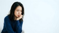 Young Asian woman with long dark hair wearing a navy blue sweater, resting her face on her hand with a contemplative expression against a white background.