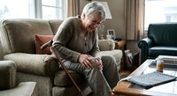 Senior Caucasian man with gray hair sitting on beige sofa holding walking cane in bright living room with natural light from windows for healthcare concepts.