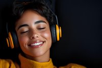 Young mixed race woman with freckles wearing yellow turtleneck sweater and orange headphones smiling with eyes closed against dark background, expressing joy and relaxation.