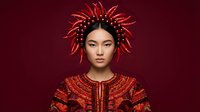 Young woman with East Asian features wearing ornate red and gold embroidered garment and elaborate spiked headdress against deep burgundy background for fashion, editorial, and artistic projects.