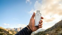 Clear water bottle held in hand against mountain landscape and sky during outdoor adventure or hiking trip.