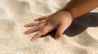 Child's hand touching soft golden sand on beach, creating sensory experience with natural texture and warm sunlight casting gentle shadows.