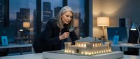 Mature woman with gray hair and glasses examining architectural model with illuminated buildings in modern office at night with city skyline view.