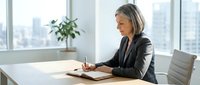 Mature woman with gray hair, Caucasian ethnicity, wearing black blazer, writing in notebook at wooden desk in modern high-rise office with city skyline view, professional business environment.