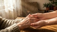 Young caregiver gently holds elderly person's wrinkled hands, expressing empathy and support in a warm, sunlit home setting.