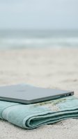 Laptop computer resting on folded light blue towel on sandy beach with ocean waves blurred in background, representing remote work, digital nomad lifestyle, and summer productivity.