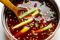 Spicy Asian broth with sesame seeds, green onions and wooden spoon in metal pot. Rich dark red soup base garnished with fresh vegetables and aromatics.