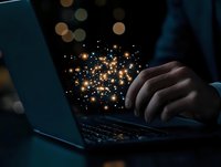 Glowing magical sparkles emanate from laptop keyboard while person types in dark room, creating ethereal atmosphere with bokeh light effects in background.