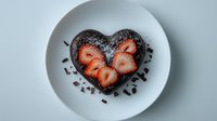 Heart-shaped chocolate dessert with fresh strawberry slices on top, garnished with chocolate sprinkles, served on white plate against light background.