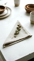 Minimalist table setting with folded linen napkin, eucalyptus sprig, and neutral ceramic dishware arranged on white surface for dining and event styling.