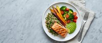 Grilled salmon fillet with quinoa, avocado slices, roasted carrots, broccoli, and cherry tomatoes on a white plate, marble background.