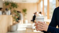 Businesswoman holding white ceramic cup in modern office with colleagues and plants in background, suitable for corporate, workplace, and beverage marketing content.