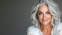 Confident mature woman with silver gray wavy hair and natural makeup wearing white lace top, smiling against dark background. Aging gracefully concept.