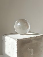 Clear glass sphere on concrete pedestal casting soft shadows in minimalist composition. Natural light creates ethereal atmosphere for modern interior design.
