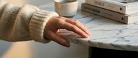 Caucasian woman's hand resting on marble desk surface near coffee cup and stacked books, wearing beige knit sweater, workspace organization and lifestyle concept.