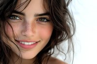 Close up portrait of young Caucasian woman with natural freckles, dark wavy hair, and bright smile showing genuine happiness and confidence against light background.
