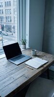 Modern workspace with laptop, notebook, and coffee mug on wooden desk by window overlooking urban cityscape. Minimalist office setup for remote work, freelancing, and productivity.
