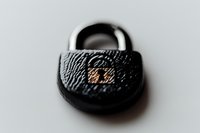 Close-up macro shot of black metal padlock with textured surface and copper-colored keyhole detail against blurred light background, symbolizing security and protection.