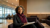 Professional African American businesswoman with curly hair wearing dark blazer and burgundy scarf sitting in modern office lobby holding documents and smiling confidently.