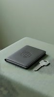 Black leather passport holder with globe emblem and silver key on sage green fabric surface, suitable for travel, security, and lifestyle design projects.
