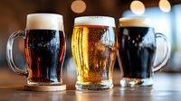 Three frosty beer mugs filled with different craft brews - light lager, dark stout and amber ale on wooden coasters against blurred pub background with bokeh lights.