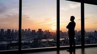Silhouette of male professional standing at floor-to-ceiling windows overlooking sprawling cityscape during golden sunset, contemplating urban landscape from high-rise office building.