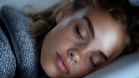 Young Caucasian woman with freckles sleeping peacefully in bed wrapped in cozy gray knit blanket. Natural morning light illuminates serene facial features.
