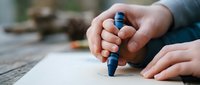 Close-up of hands holding blue crayon drawing on white paper outdoors, creative art activity for children and adults, artistic expression and learning.
