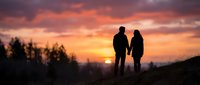 Silhouetted couple standing together on hillside at sunset with dramatic orange and golden sky, evergreen forest landscape, romantic outdoor scene for travel and lifestyle content.