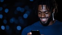 Young African American man with dreadlocks smiling while using smartphone at night, blue bokeh lights in background creating modern urban atmosphere.