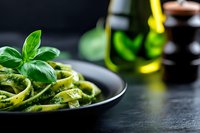 Fresh green pasta with basil pesto sauce served in black bowl on dark wooden table with olive oil bottles in background for Italian cuisine recipes.
